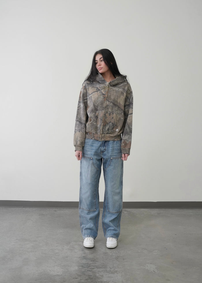 A female model wearing an Akimbo style camo jacket from Obscure Collective, blending modern streetwear aesthetics with a relaxed fit. The hoodie features a minimalist design, subtly loose sleeves, and soft knitted fabric, offering both comfort and style in a neutral tone that's perfect for versatile layering.